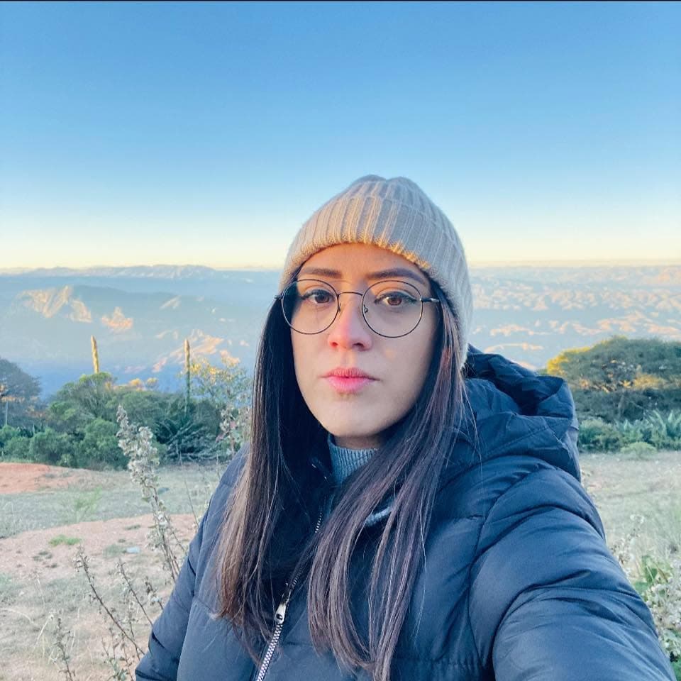 Mujer con gafas y gorro posando frente a un paisaje montañoso durante la hora dorada.