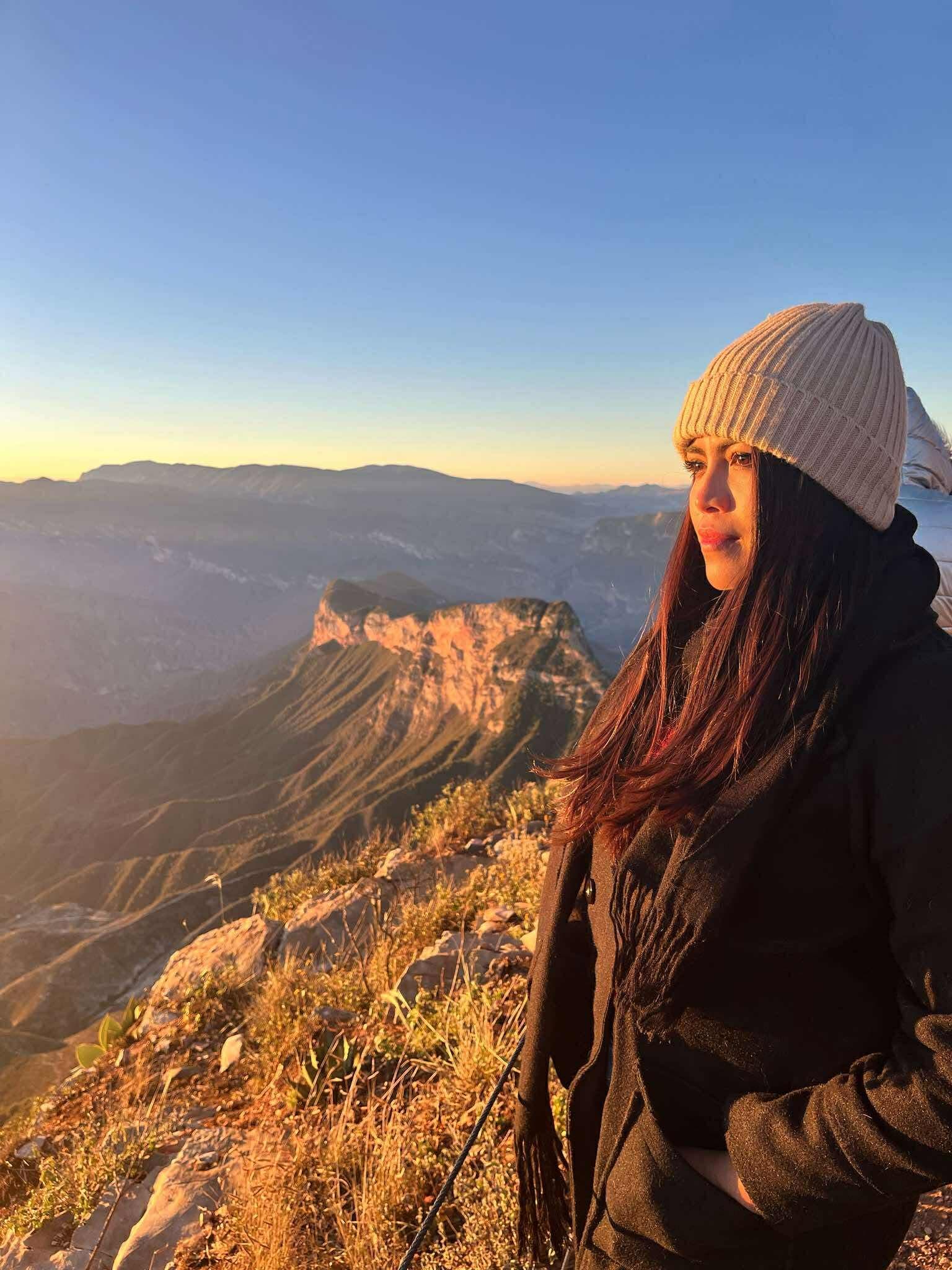 Mujer con gorro beige observa un vasto cañón montañoso bajo la cálida luz del atardecer.