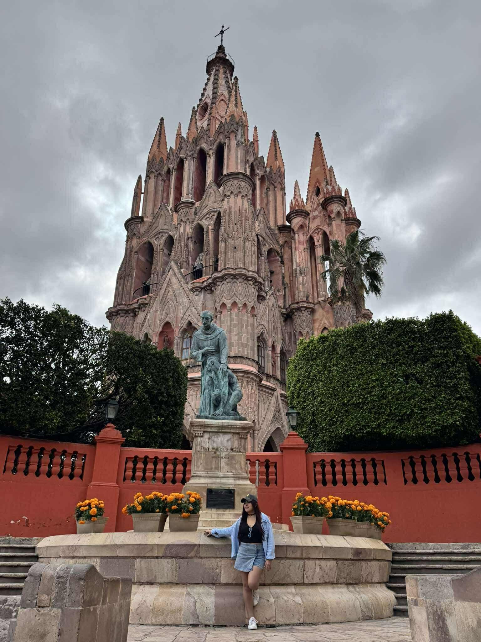 Mujer posando frente a la Parroquia de San Miguel Arcángel en un día nublado.
