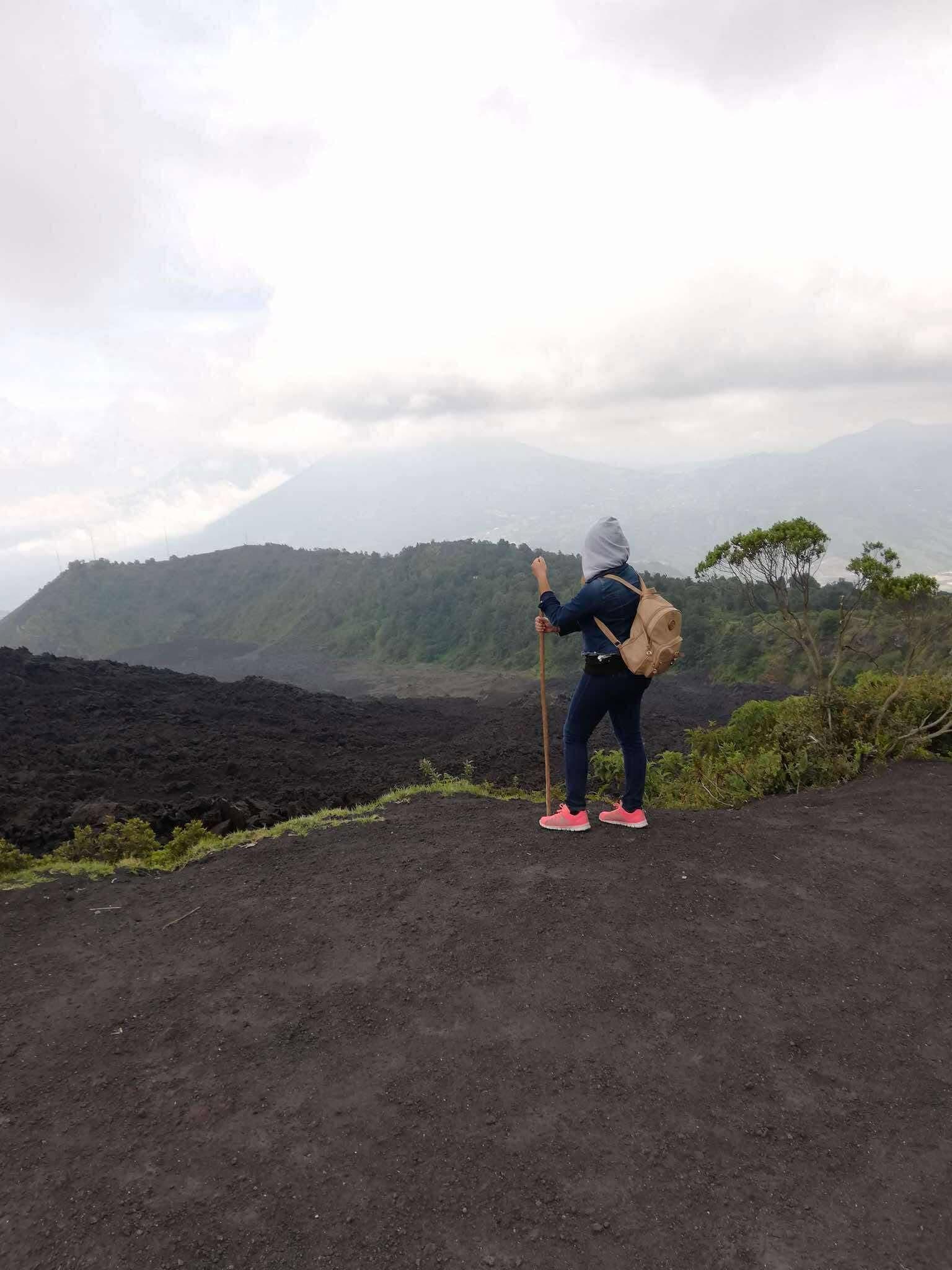 Excursionista con mochila y bastón contempla un campo de lava negra y montañas lejanas.