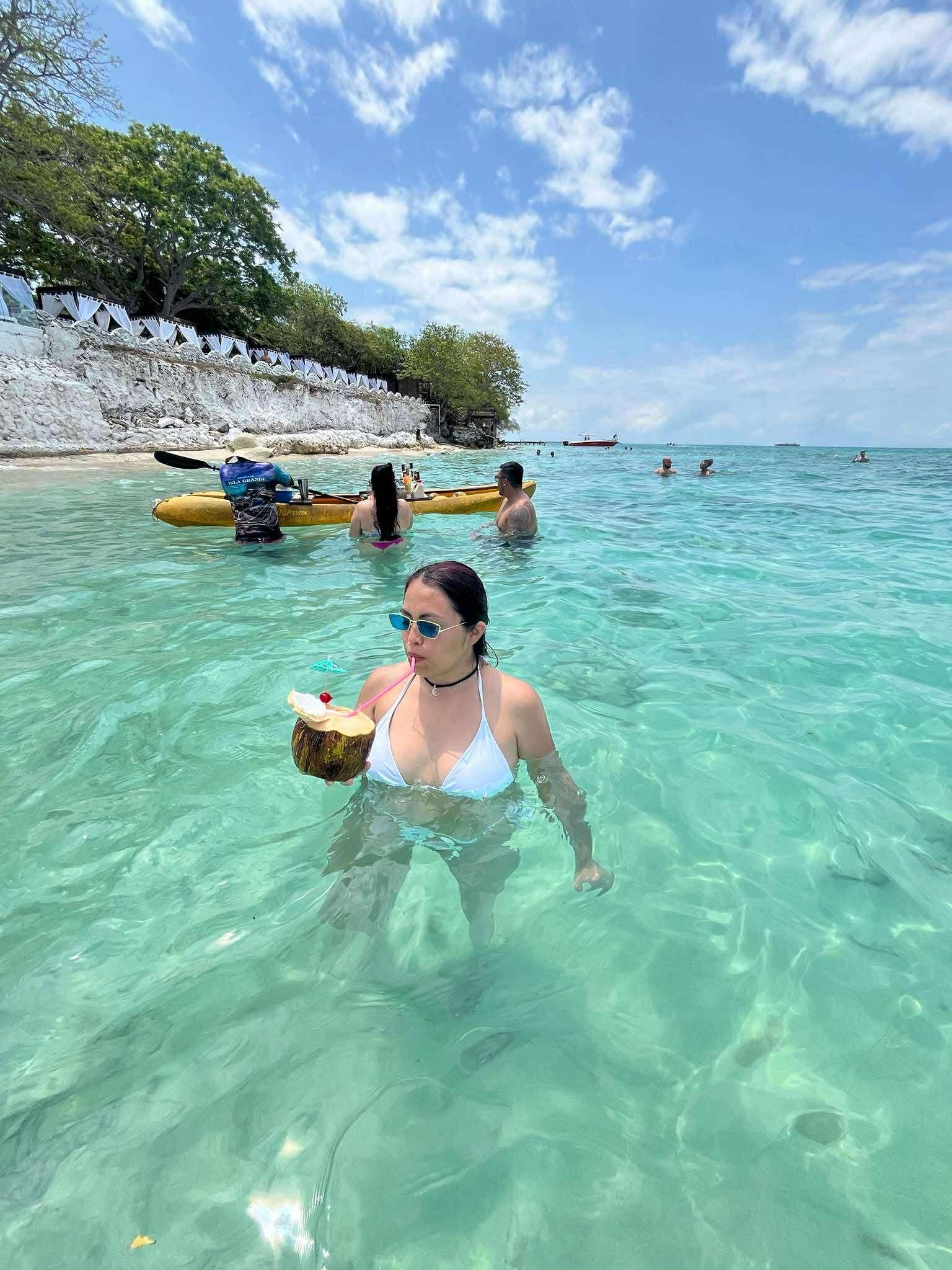 Mujer bebiendo de un coco en aguas cristalinas con un kayak amarillo al fondo.