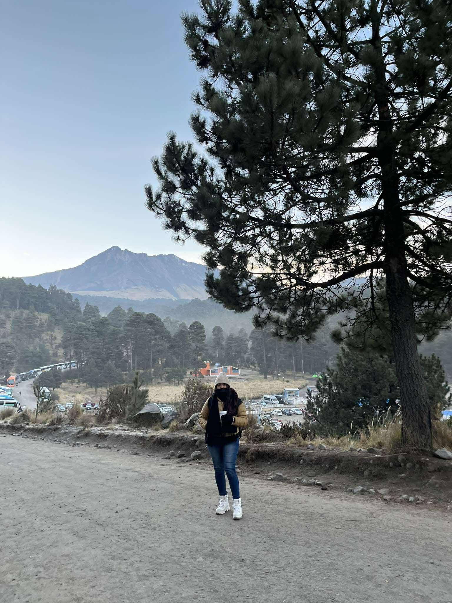 Mujer abrigada en un camino de tierra con una gran montaña y pinos al fondo.