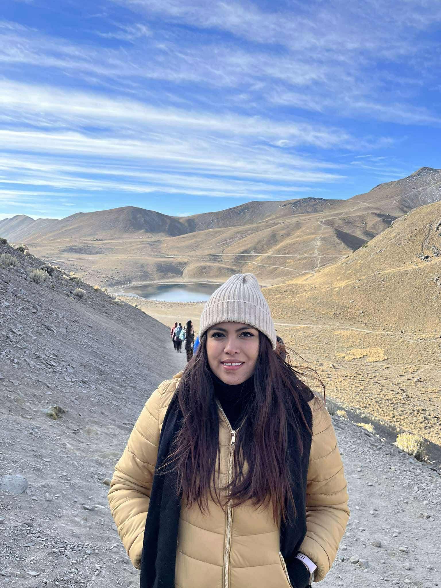 Mujer sonriente con chaqueta beige y gorro en un sendero montañoso frente a un lago.