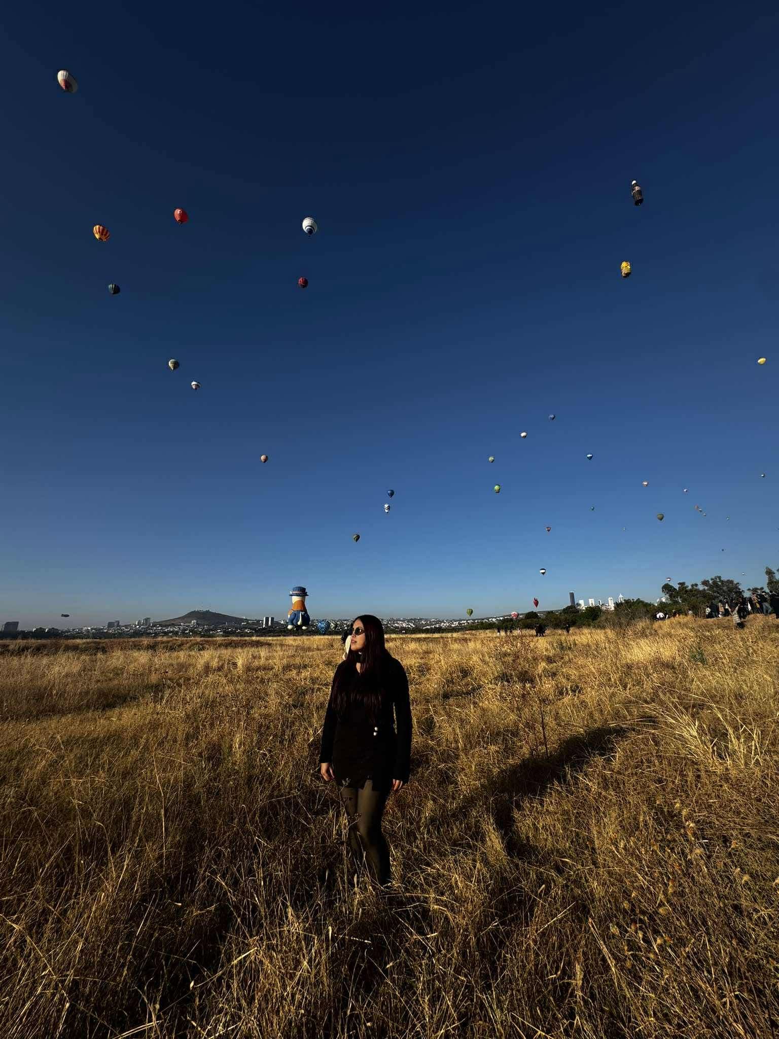 Mujer en un campo dorado bajo un cielo azul lleno de globos aerostáticos coloridos.