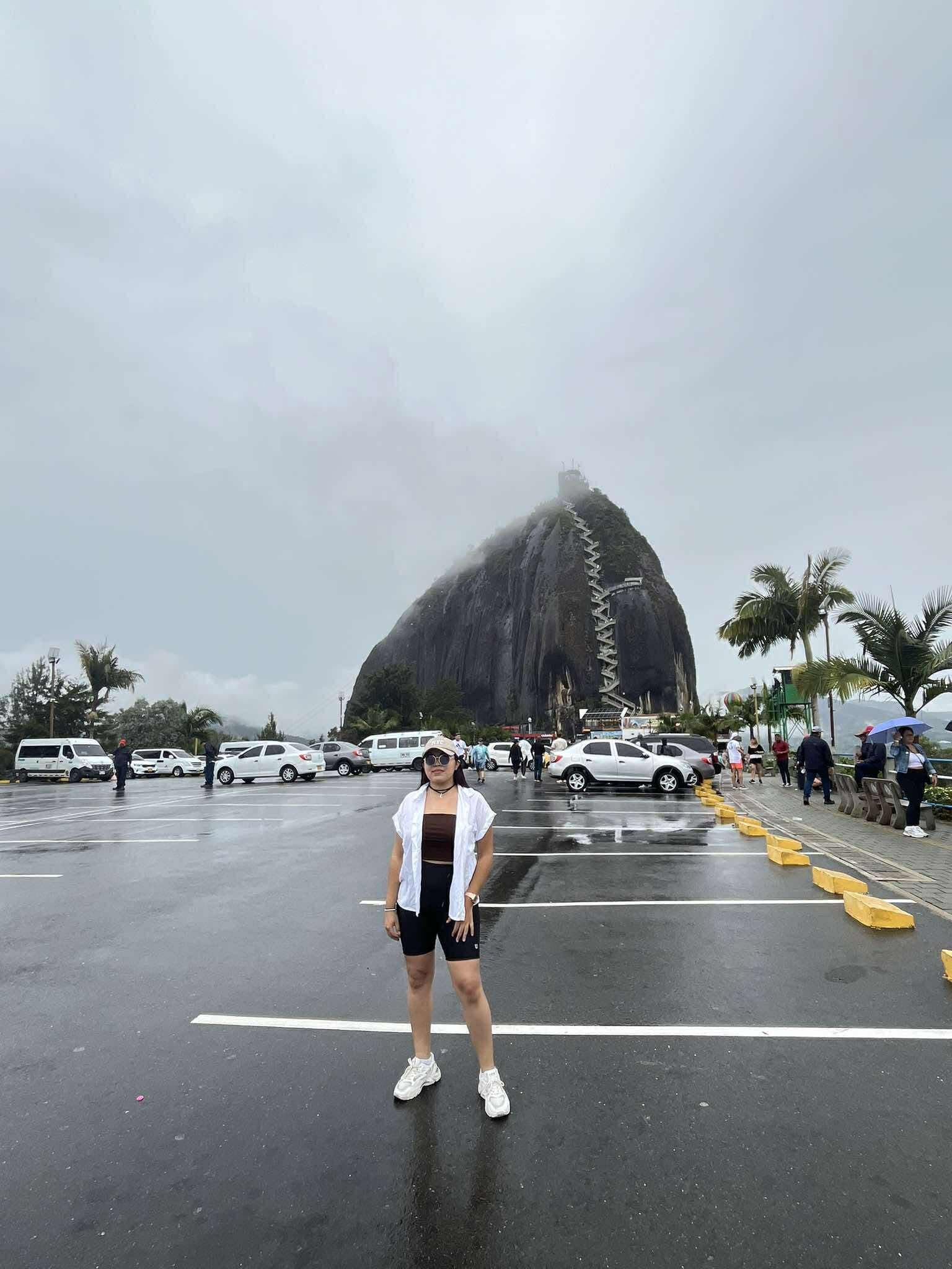 Mujer posando frente a la Piedra del Peñol en Guatapé bajo un cielo nublado.